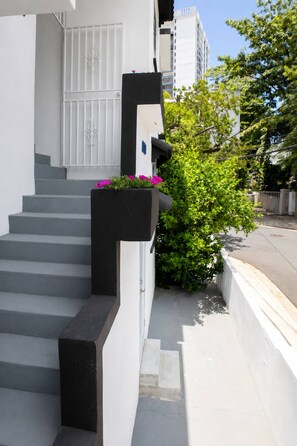 Eine Treppe führt zu einem Hauseingang, ein Blumenbeet und ein Baum befinden sich daneben.
