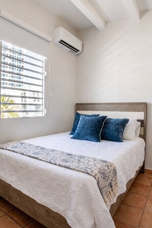 Ein Schlafzimmer mit einem Bett, einem Fenster mit Jalousien, einer Klimaanlage und Blick auf Gebäude.