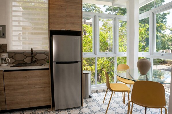 Eine moderne Küche mit Edelstahl-Kühlschrank, Holzschränken, einem Glastisch und Stühlen, mit großen Fenstern und Blick auf Grünflächen.