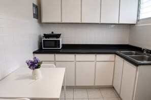 Eine kleine Küche mit weißen Schränken, einer schwarzen Arbeitsplatte, einer Mikrowelle, einem Spülbecken und einer Tischvase mit violetten Blumen.