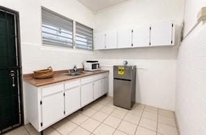 Eine kompakte Küche mit weißen Schränken, einem Edelstahl-Kühlschrank, einer Mikrowelle, einem Spülbecken und einem Fenster mit Jalousien.