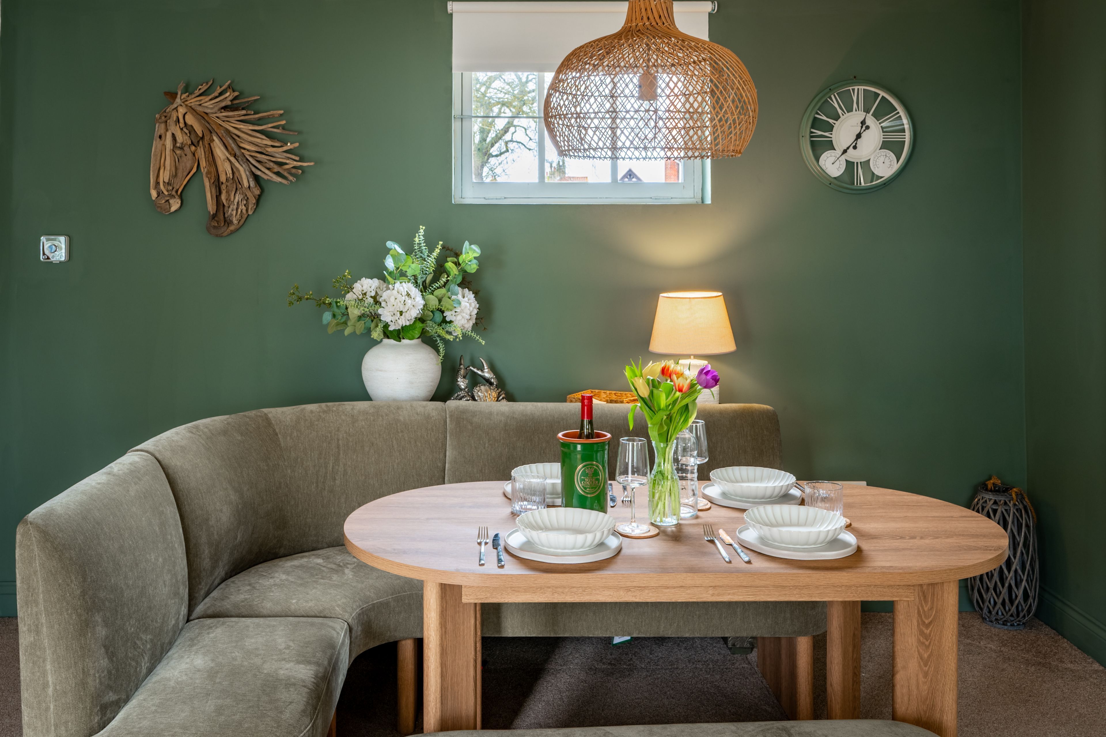 Un coin repas avec une table ronde en bois, des murs verts, un luminaire en fibres naturelles, une horloge et une fenêtre laissant entrer la lumière naturelle.