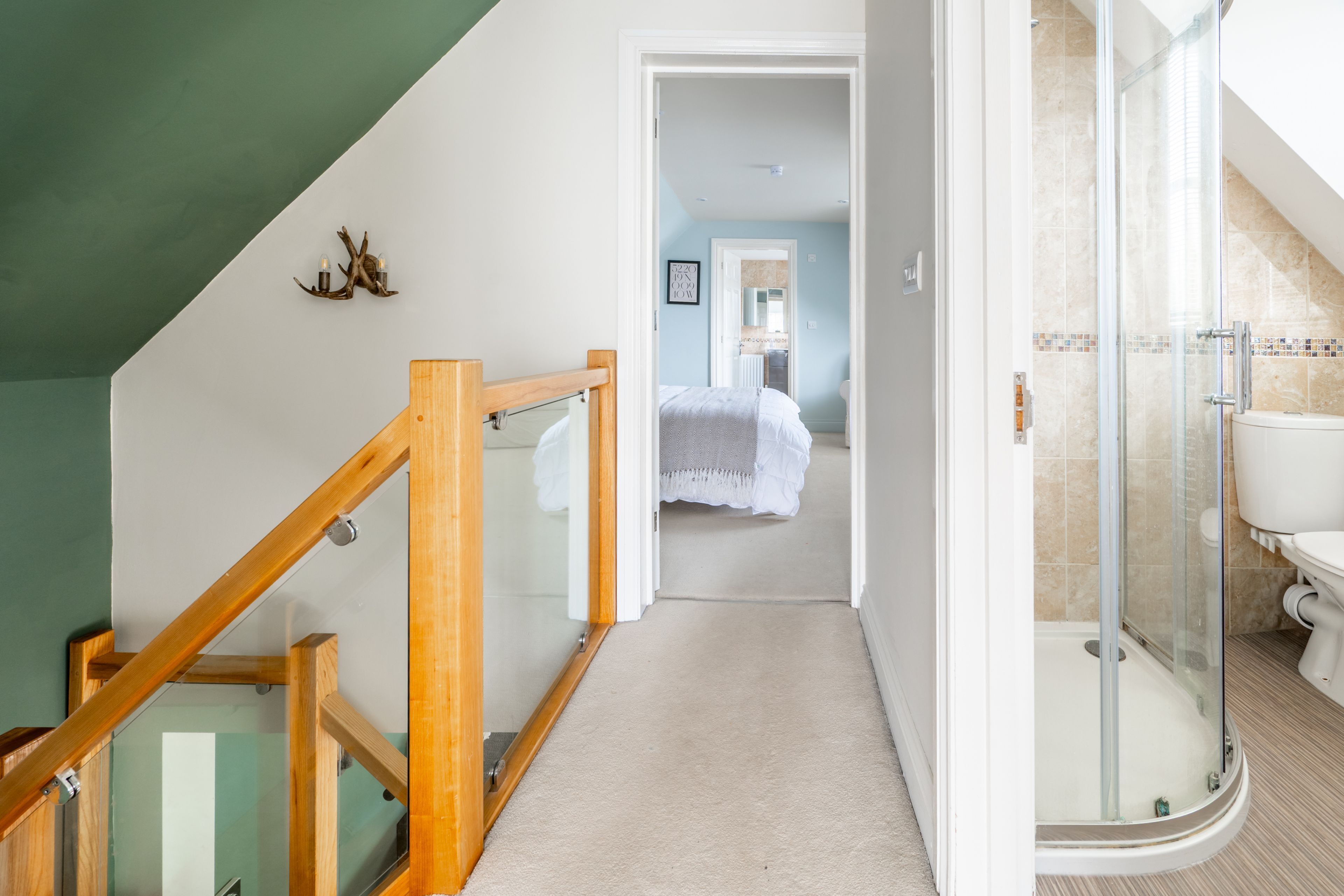 Un couloir mène à une salle de bain dotée d’une douche en verre, à un escalier en bois et à une chambre visible avec un lit.