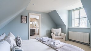 Camera da letto con un letto, cuscini, una sedia, una finestra e un ingresso che dà su un bagno.