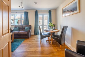 A living room with a brown leather sofa, a dining table with chairs, a green rug, and a window with curtains. - Seabreeze, Wells-next-the-Sea, Norfolk (Wells-next-the-Sea)