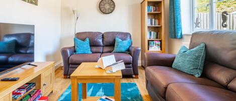 A living room with two brown leather sofas, a wooden coffee table, a bookshelf, and a window with curtains.