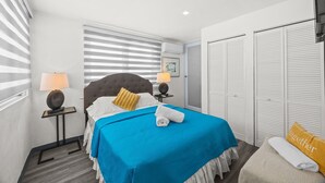 A hotel room with a bed, two bedside tables, a window with blinds, a wardrobe, and a wall-mounted air conditioner.