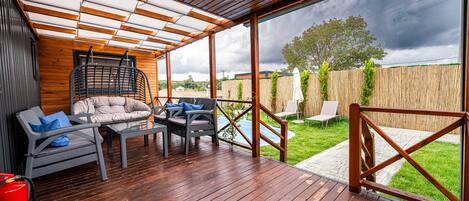 Un patio exterior techado con piso de madera, un sillón colgante y dos sillones de descanso.
