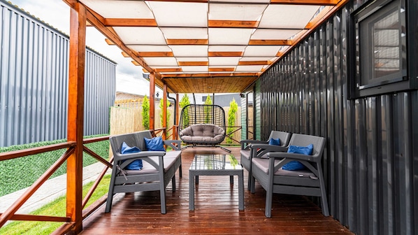 A covered outdoor seating area with a table and two chairs, a hanging chair, and a view of a garden.