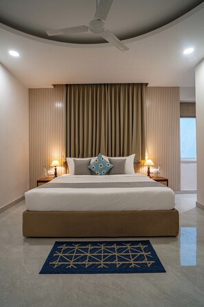 Camera d'albergo con un letto grande, una ventola a soffitto, due lampade da comodino, una finestra con tende e un tappeto blu.