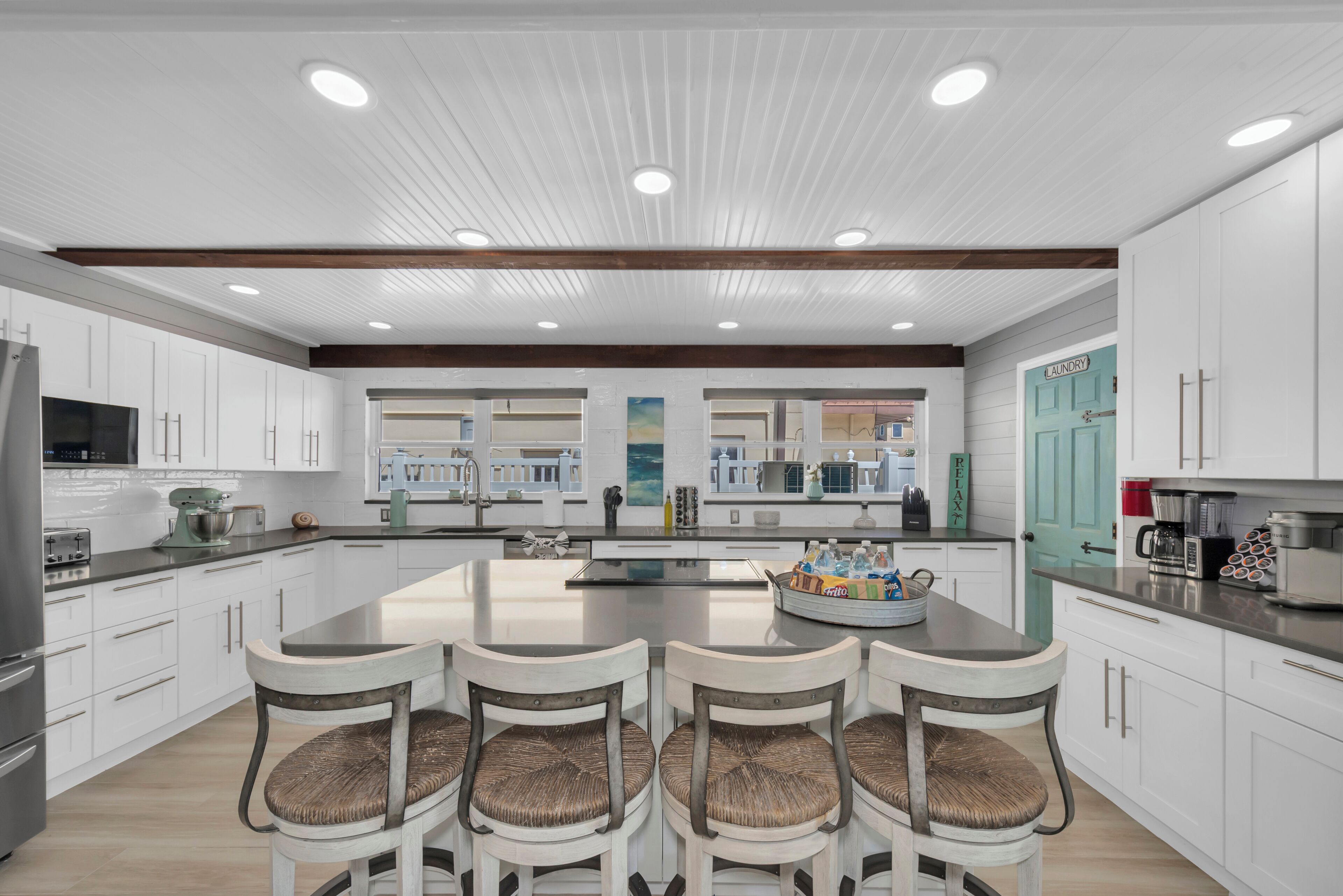 Une cuisine moderne avec des meubles blancs, un îlot central doté de tabourets de bar et une porte turquoise portant l’inscription « Buanderie ».