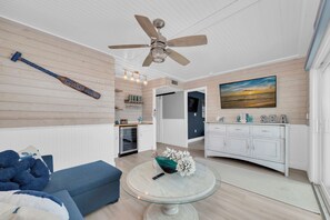 A modern living room with a blue sofa, a glass coffee table, a ceiling fan, and a kitchen area with a white cabinet and a painting of a sunset. - Beachy 4 Bedroom 3 Bath Pool Home Mins to Redington Beach (Redington Beach)