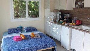 A bedroom with a bed, pillows, and a window. There is a kitchenette with a sink, stove, and cabinets. - Charming air-conditioned studio with pool between Aix-en-Provence and Marseille (Les Pennes-Mirabeau)
