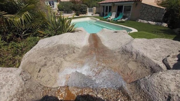 A swimming pool with a rock feature, surrounded by a well-maintained lawn and a house with a tiled roof. - Charming air-conditioned studio with pool between Aix-en-Provence and Marseille (Les Pennes-Mirabeau)