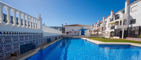 Una piscina con borde de baldosas blancas, una valla blanca con detalles decorativos, y un edificio con tejado de tejas y balcones.
