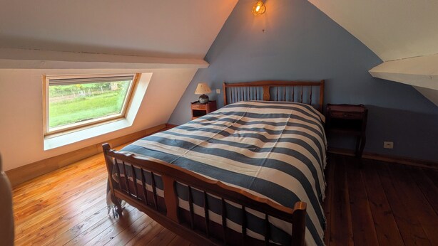 A bedroom with a sloped ceiling, a wooden bed, a nightstand, a lamp, and a window with a view of greenery. - The Otter Cottage! Vacation Rental with Indoor Pool; Chancelade Pond 500 meters away (Charensat)