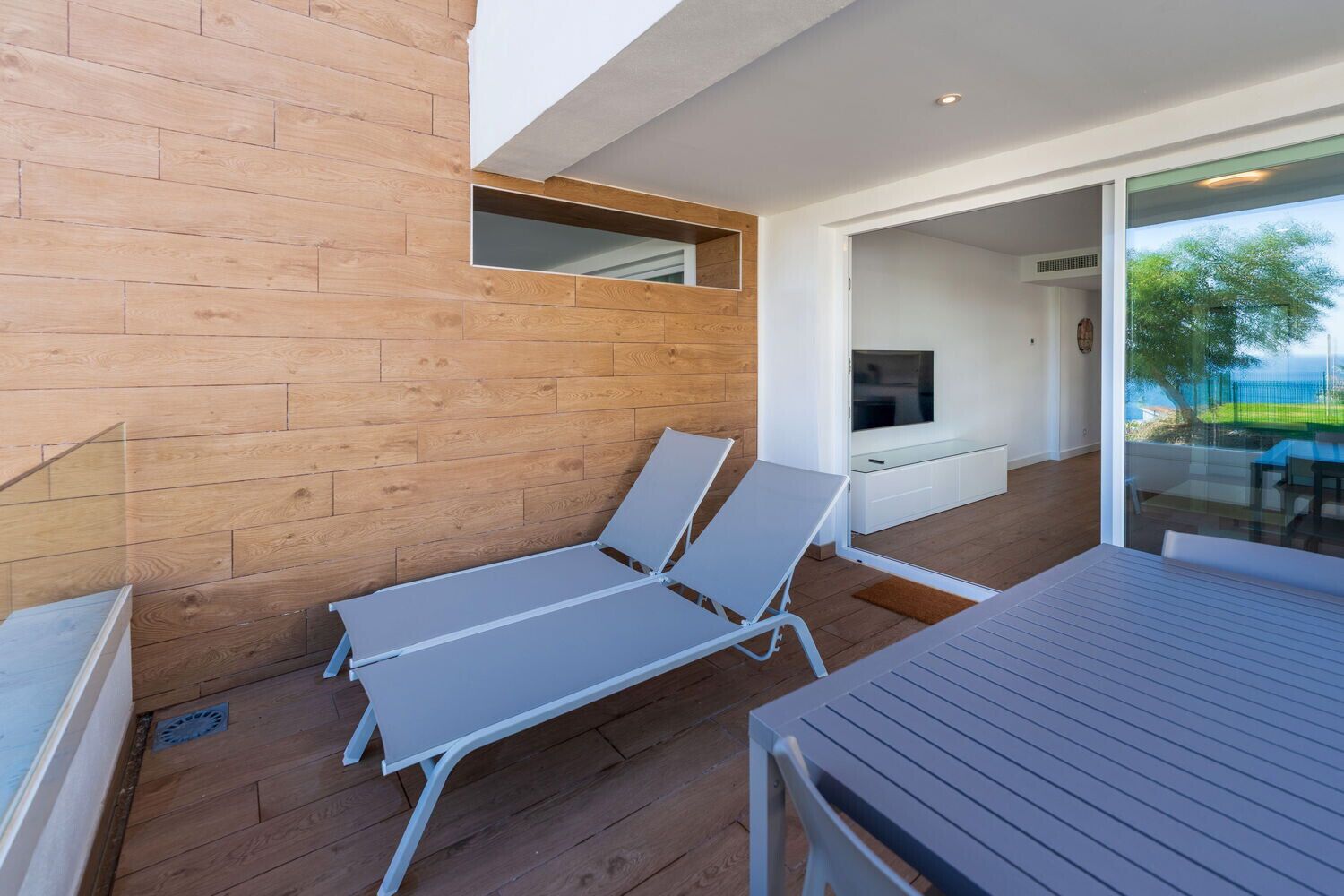 A modern outdoor lounge area with wooden flooring, two white lounge chairs, a glass table, and a TV mounted on the wall.