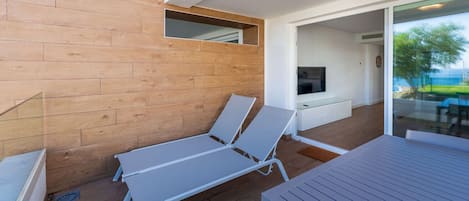Uma área de estar moderna ao ar livre com piso de madeira, duas poltronas brancas, uma mesa de vidro e uma TV fixada na parede.