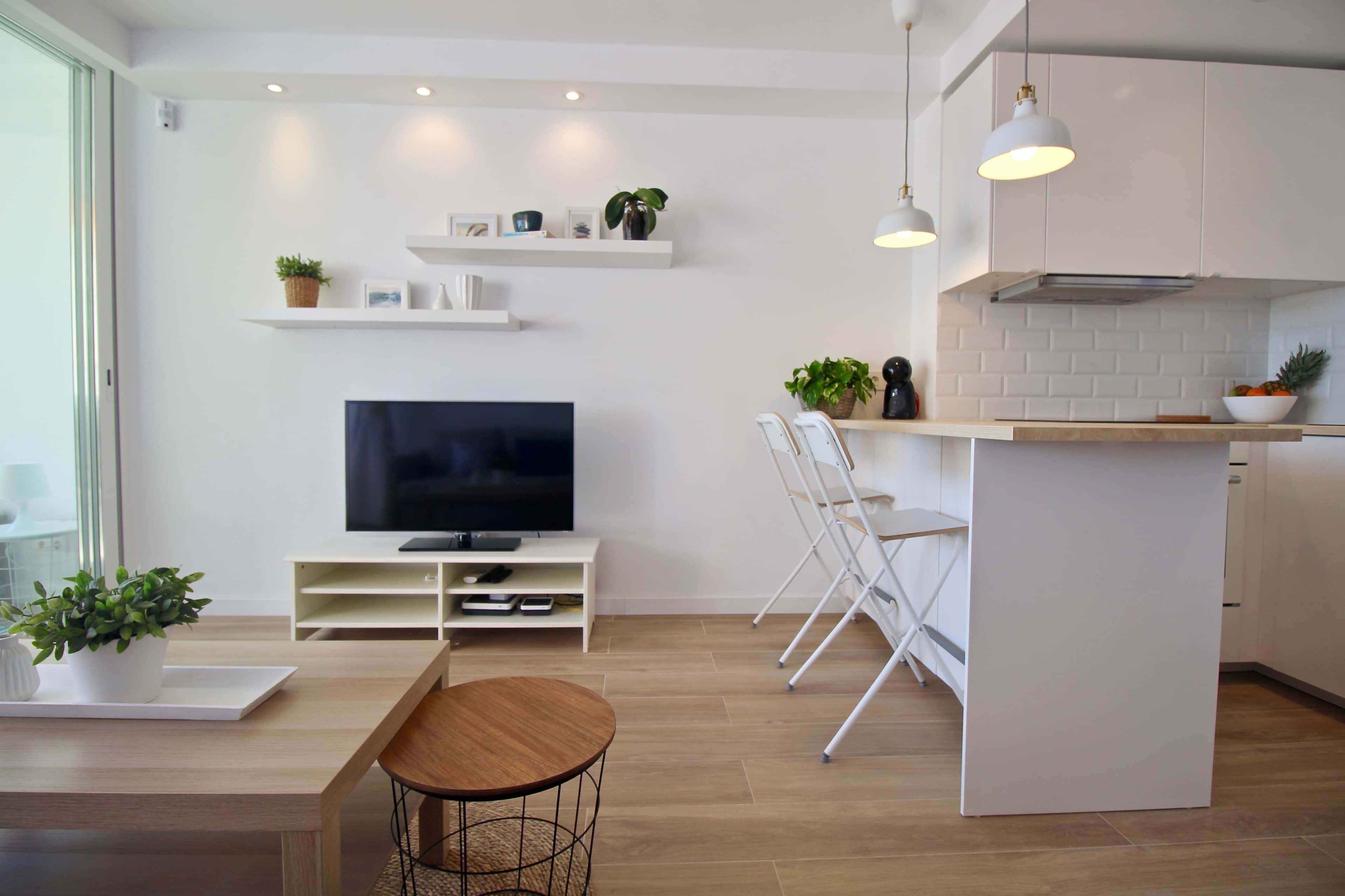 Une cuisine moderne avec des meubles blancs, un îlot central, un coin repas avec une table en bois et un téléviseur posé sur un meuble blanc.