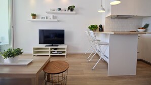 Et moderne køkken med hvide skabe, en central ø, et spiseområde med et træbord og et fjernsyn på et hvidt stativ.