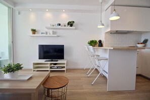 Eine moderne Küche mit weißen Schränken, einer zentralen Kochinsel, einem Essbereich mit Holztisch und einem Fernseher auf einem weißen Lowboard.