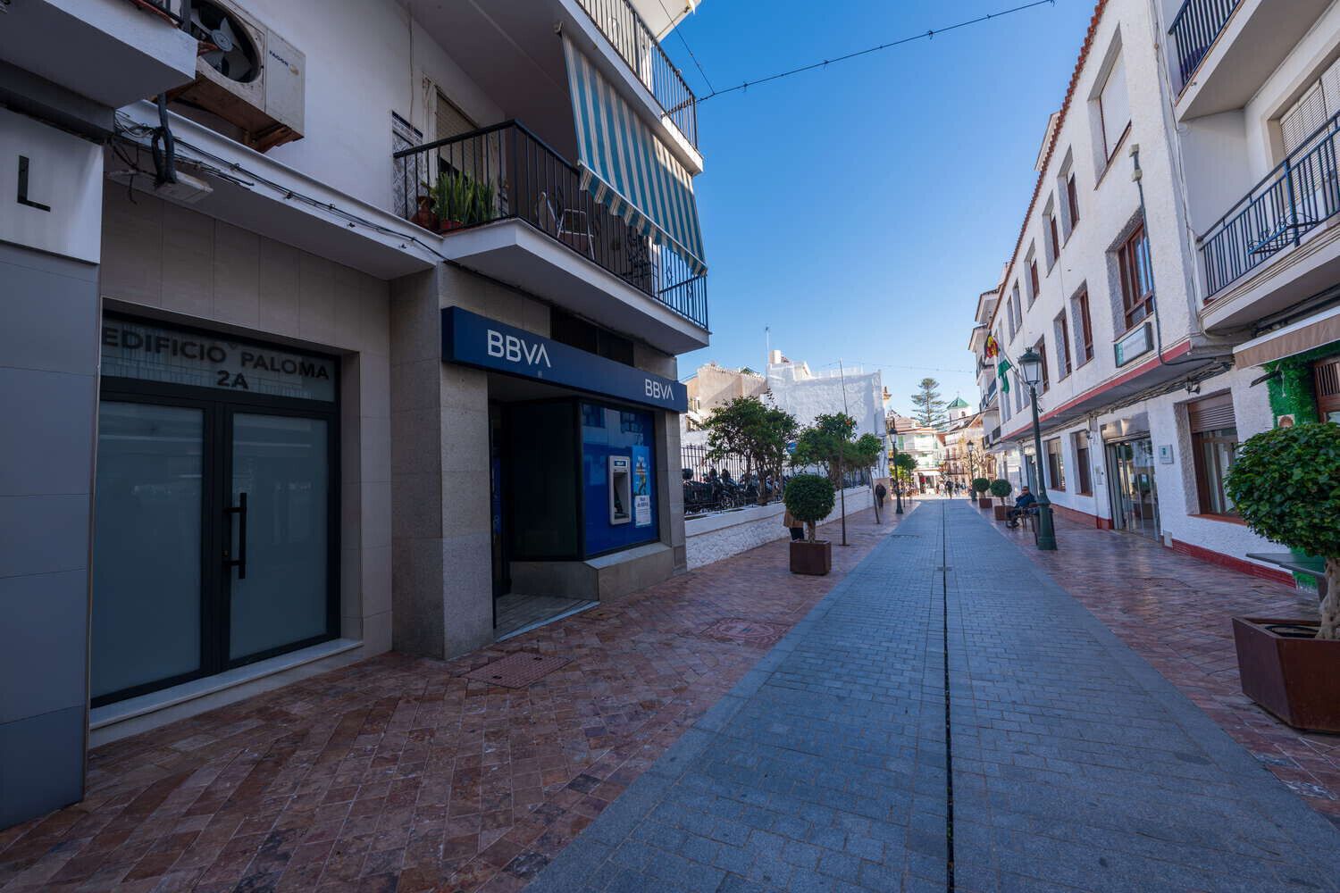 Una calle estrecha flanqueada por edificios, con una sucursal del banco BBVA y un edificio identificado como 'EDIFICIO PALOMA 2A'.