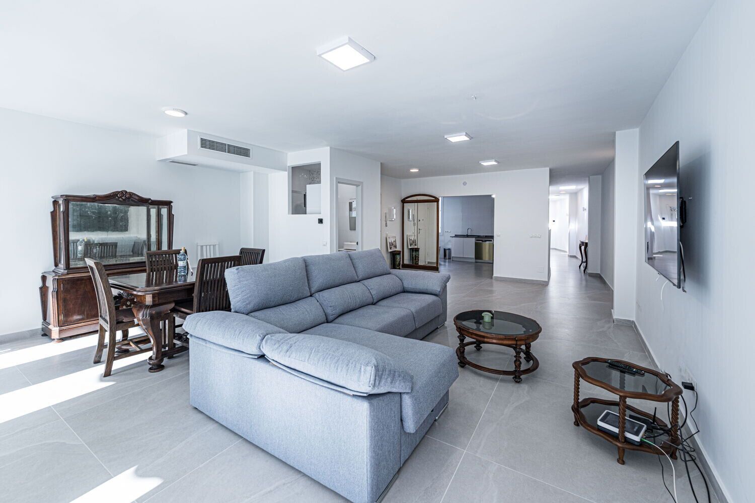 Ein modernes Wohnzimmer mit einem grauen Ecksofa, einem Holzesstisch mit Stühlen, einem Glastisch und einem an der Wand befestigten Fernseher.