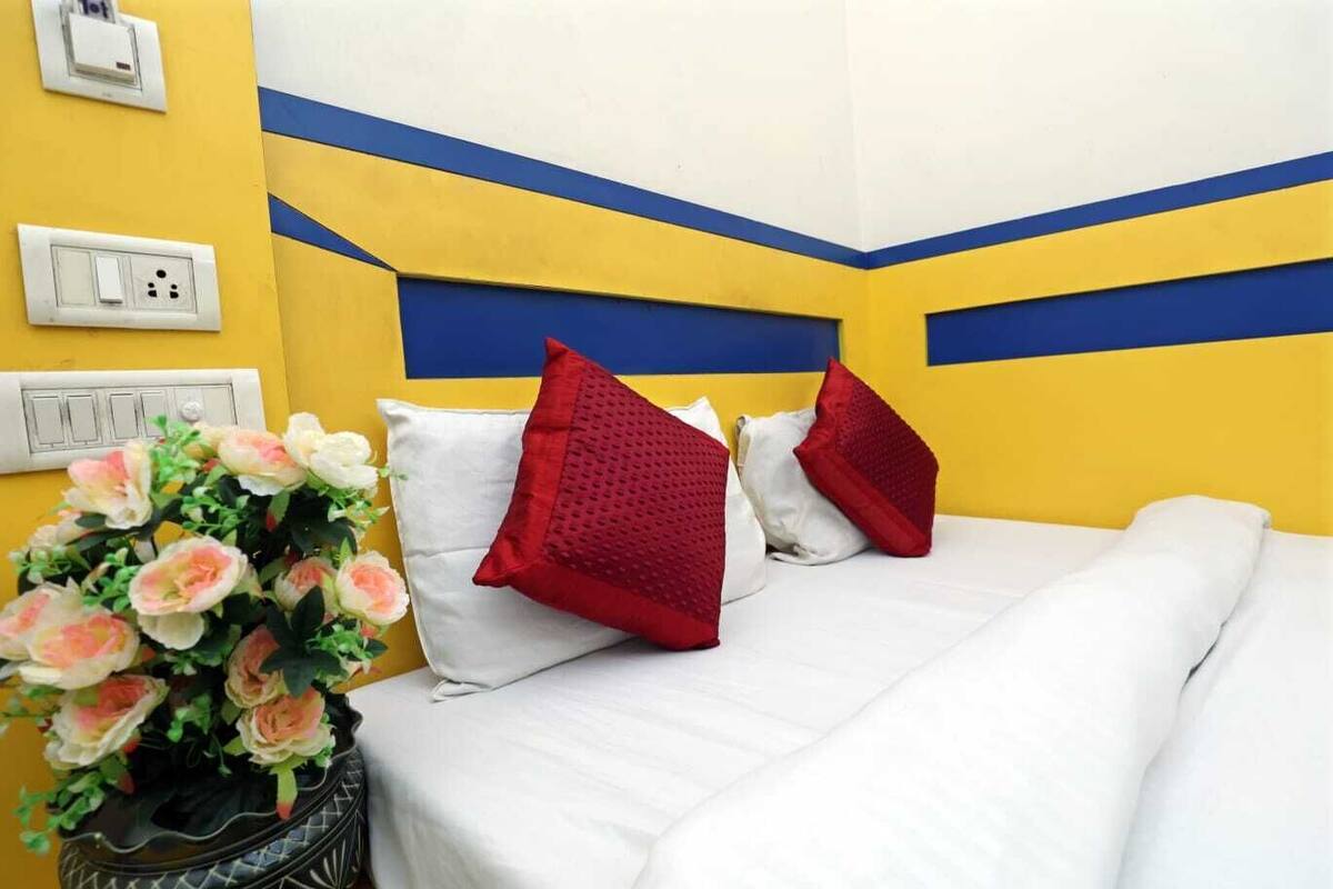 A hotel room with a bed, white bedding, red pillows, a vase of flowers, and a yellow and blue wall design.
