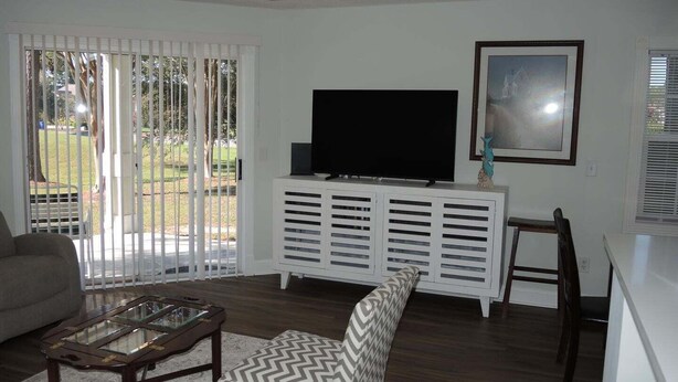 Uma sala de estar com televisão, um móvel branco para entretenimento, uma mesa de centro de vidro, uma poltrona estampada e uma mesinha de madeira.