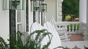 Varanda com móveis de exterior brancos, um cesto com samambaia e duas lanternas fixadas na parede.