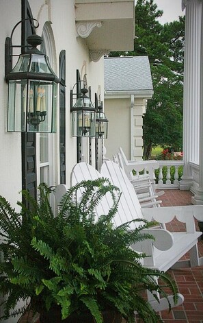 Eine Veranda mit weißen Gartenmöbeln, einem Topf-Farn und zwei wandmontierten Laternen.