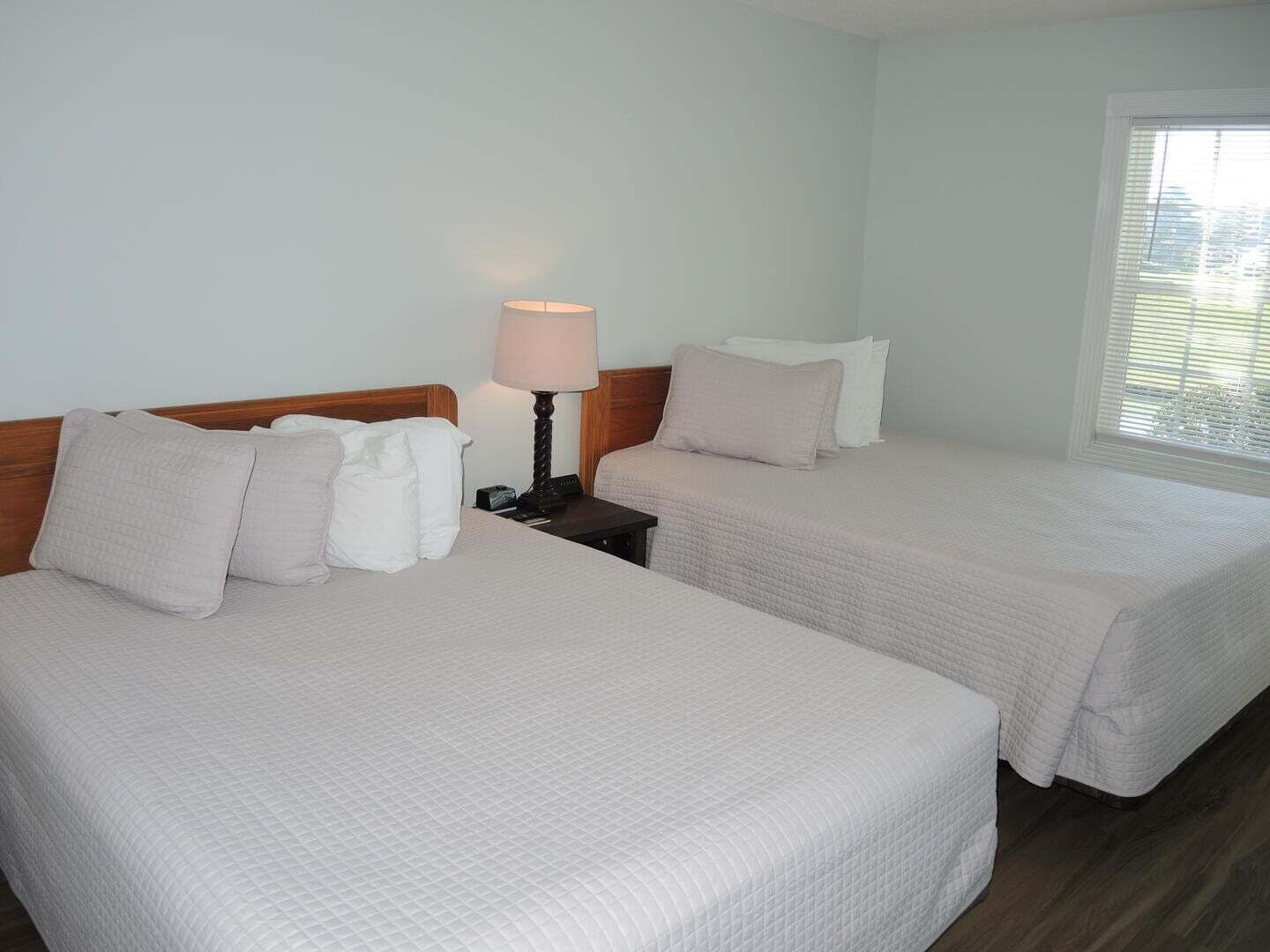 A hotel room with two beds, a bedside table with a lamp, and a window with blinds.