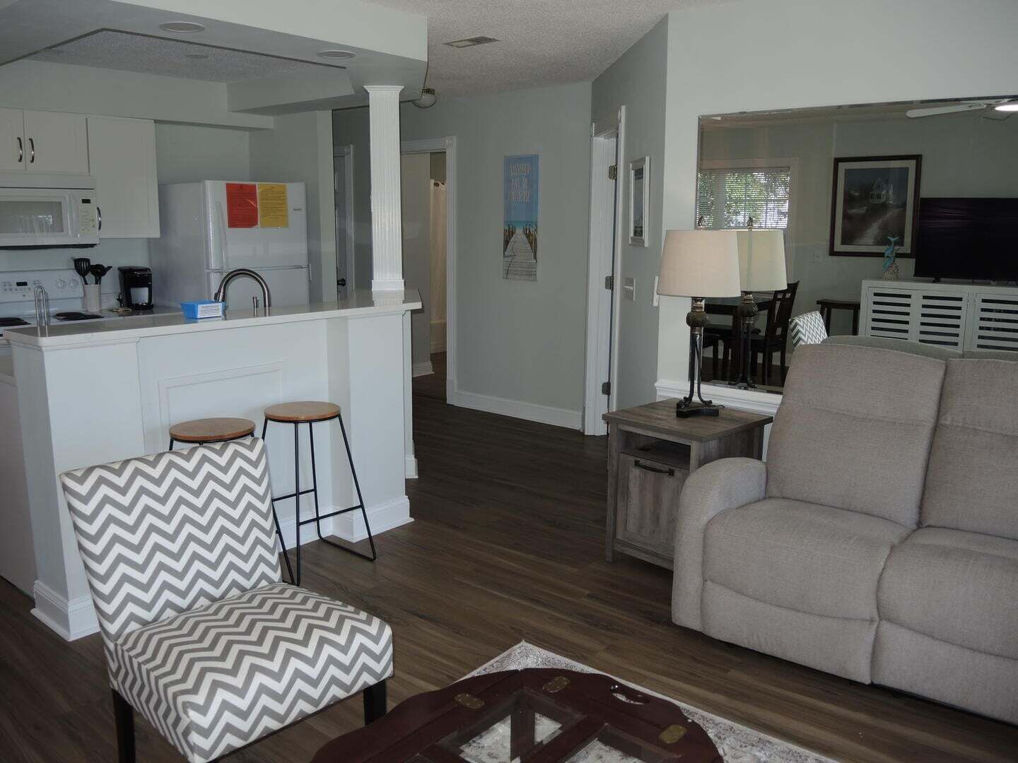 A living room with a sofa, a chair, a small table with a lamp, and a kitchen area with a microwave and a refrigerator.