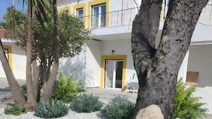 Un edificio con puerta amarilla, un balcón con barandilla metálica y un jardín con plantas y rocas.