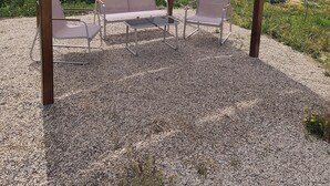 Un espacio de estar al aire libre con techo, con dos sillas y una mesa, rodeado de grava y vegetación.