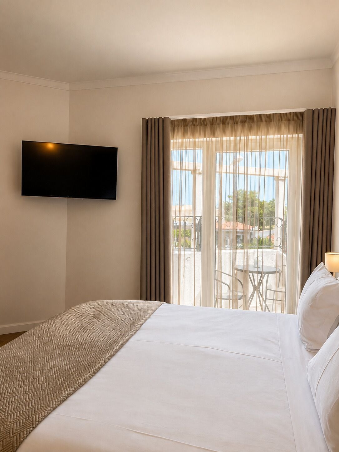 Habitación de hotel con cama, televisor de pantalla plana en la pared, balcón con mesa y sillas, y vistas al exterior.