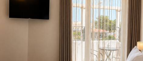 Habitación de hotel con cama, televisor de pantalla plana en la pared, balcón con mesa y sillas, y vistas al exterior.