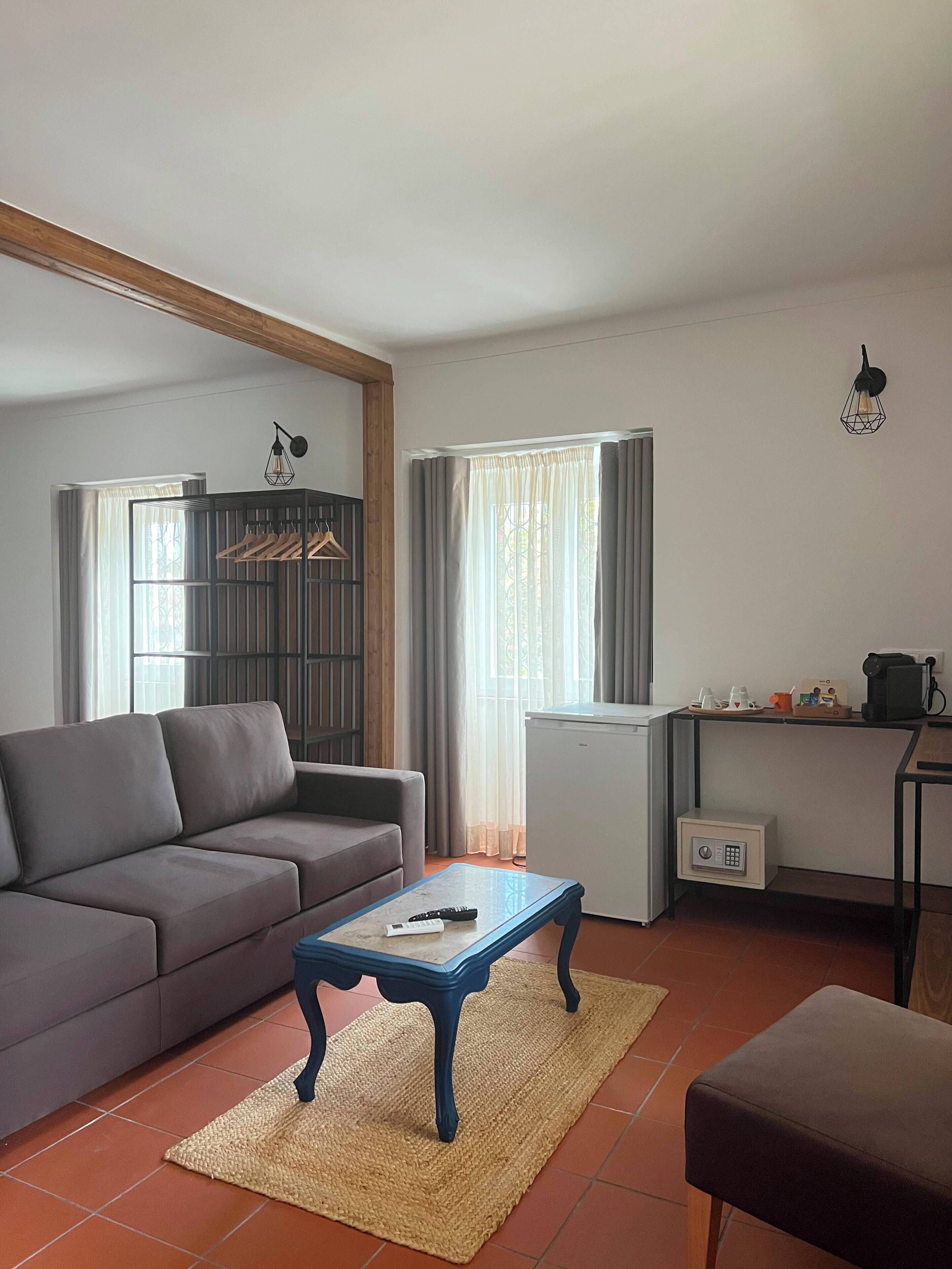 Una sala de estar con sofá, una mesa de centro, una mesita con hervidor y un refrigerador.
