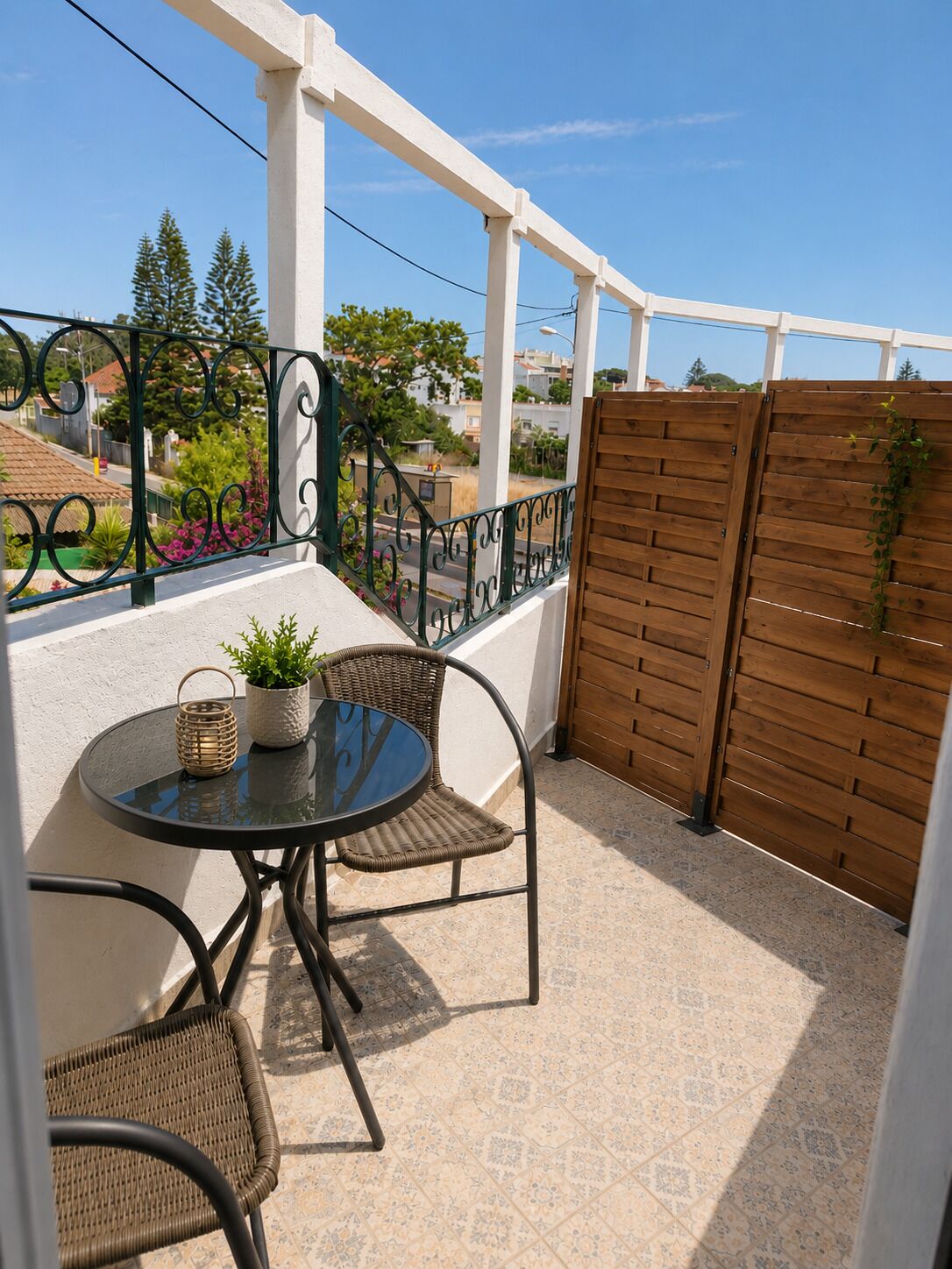 Een balkon met een ronde tafel en twee rieten stoelen, een houten hek en uitzicht op de omgeving.