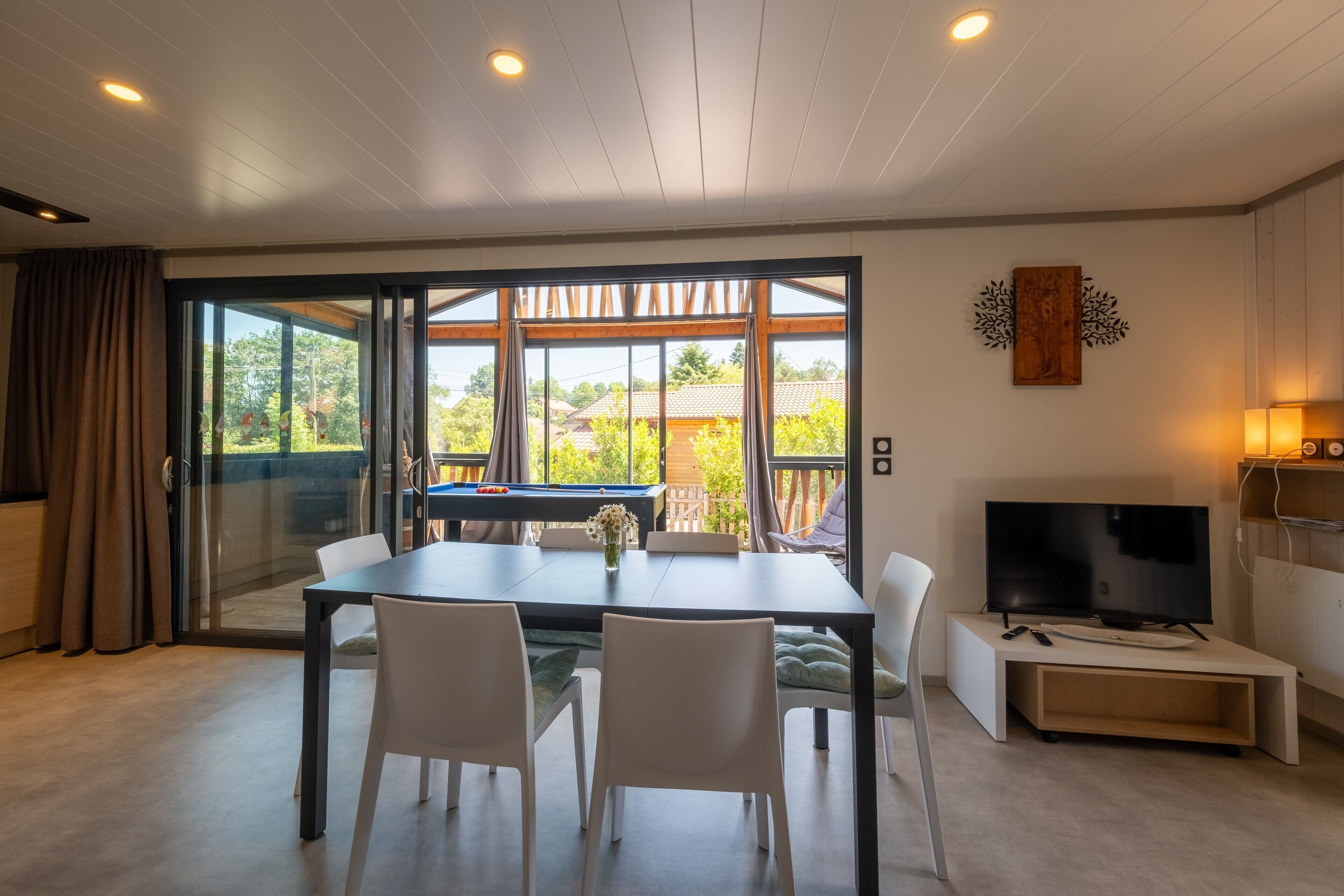 A modern living room with a dining table, chairs, a TV, and a large window with curtains.