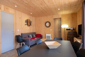 Ein Zimmer mit Holzwänden, einer Couch, Sesseln, einem Tisch, einer Lampe und einem Fernseher.