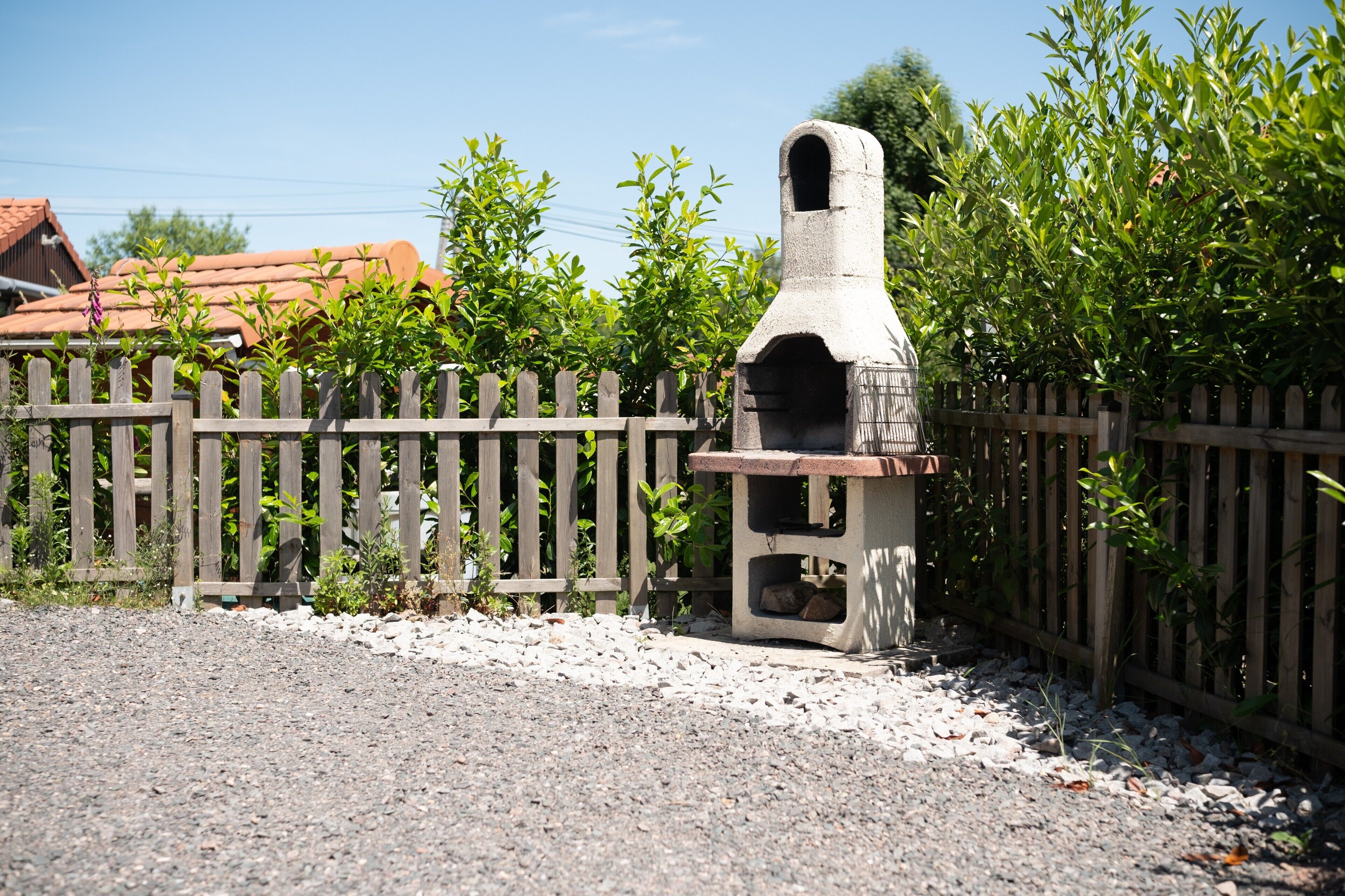 Une structure de four extérieur en pierre, dotée d’une cheminée, entourée d’une clôture en bois et bordée d’un sol en gravier.