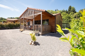 A two-story wooden cabin with a balcony, surrounded by gravel and greenery.