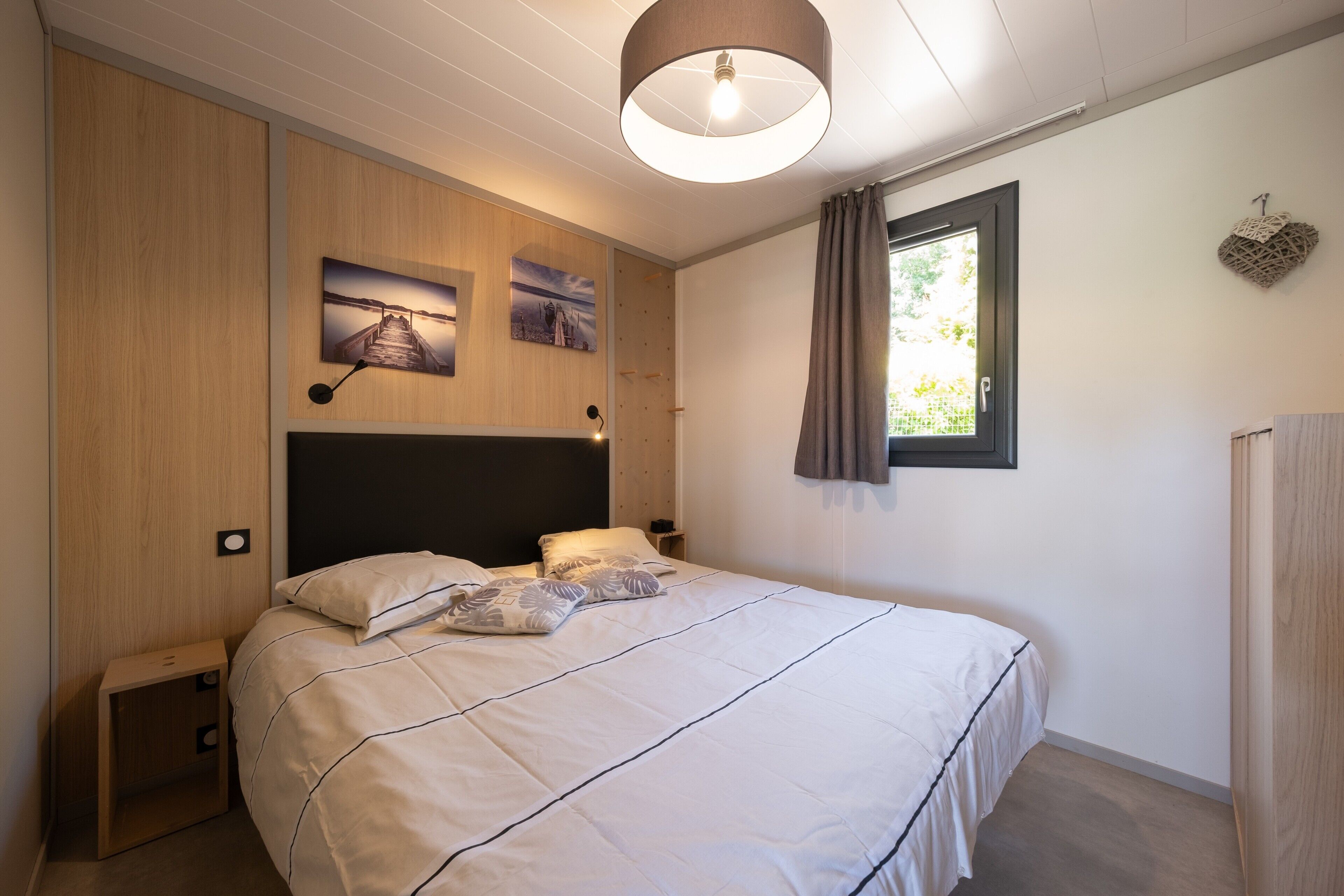 Une chambre à coucher avec un lit, un lambris en bois, une tête de lit, une lampe fixée au mur, une fenêtre avec des rideaux et une petite table de chevet.