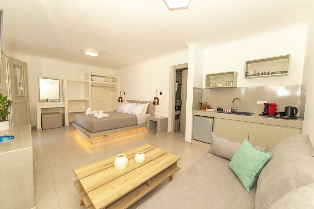 A modern hotel room with a bed, a wooden table with vases, a sofa with a green pillow, a kitchenette, and a bathroom visible through an open door.