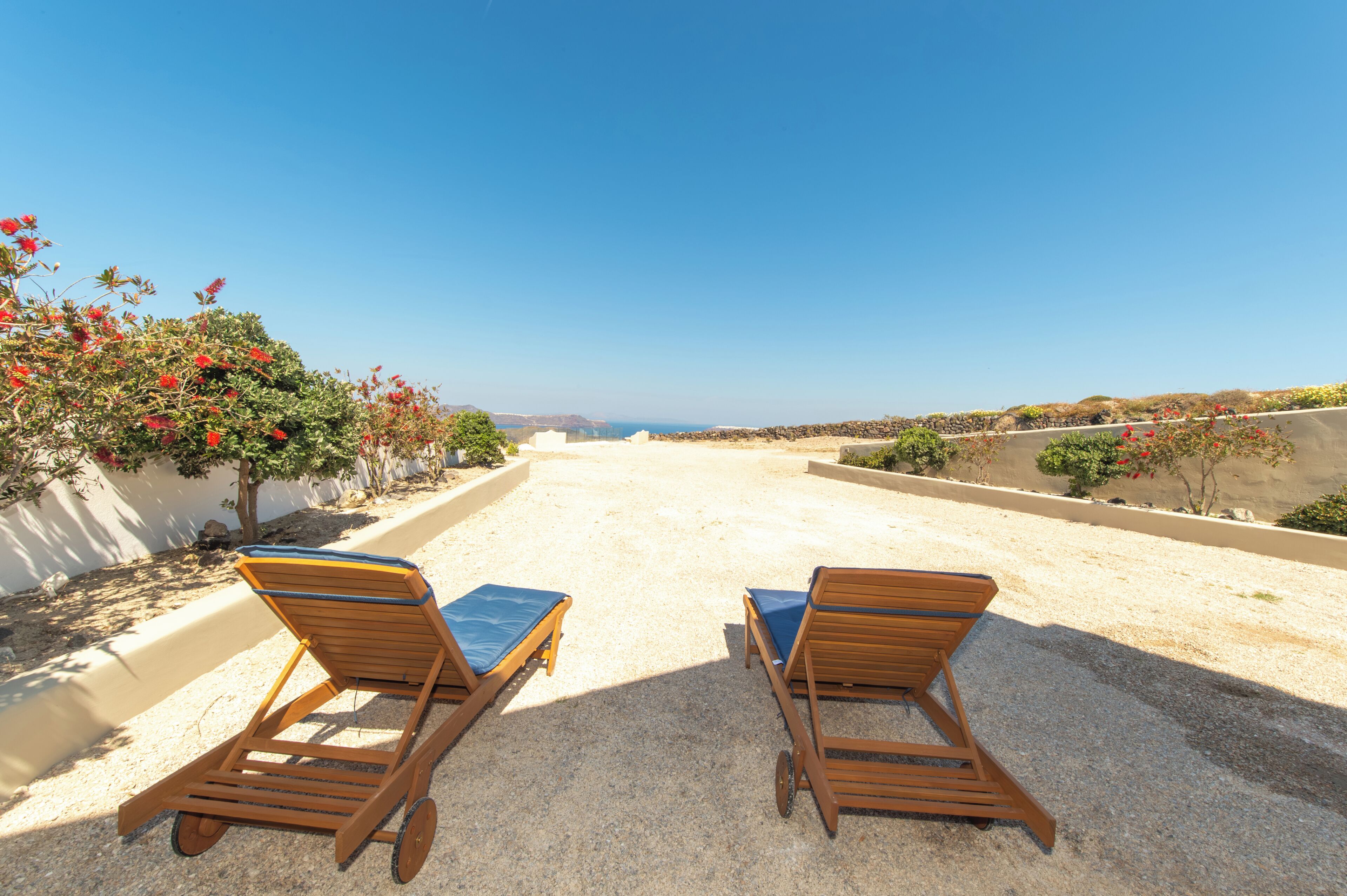 Two wooden lounge chairs with blue cushions on a gravel patio with a view of the sea.