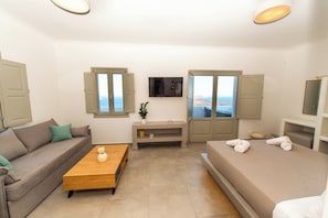 Ein Zimmer mit einer Couch, einem Bett, einem Holztischchen, einem Fernseher und Blick aufs Meer.