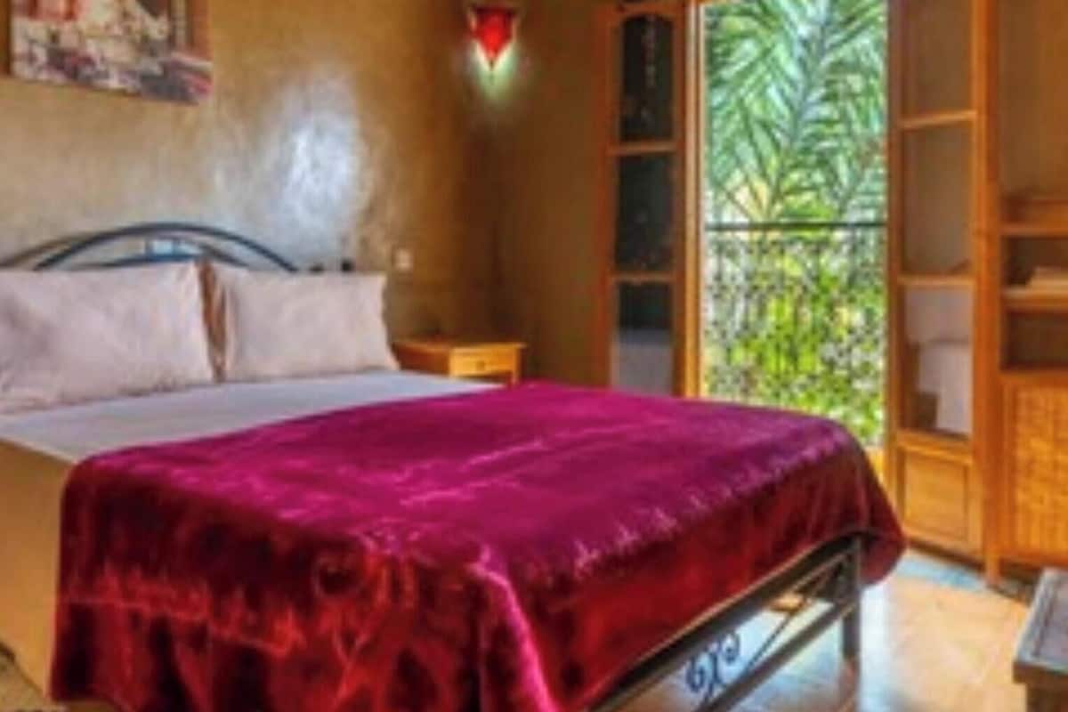 A bedroom with a bed, a nightstand, a window with a view of greenery, and a wall-mounted air conditioner.