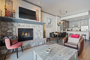 Ein Wohnzimmer mit Kamin aus Stein, einem Flachbildfernseher, einem Essbereich und einer Küche mit grauen Schränken.