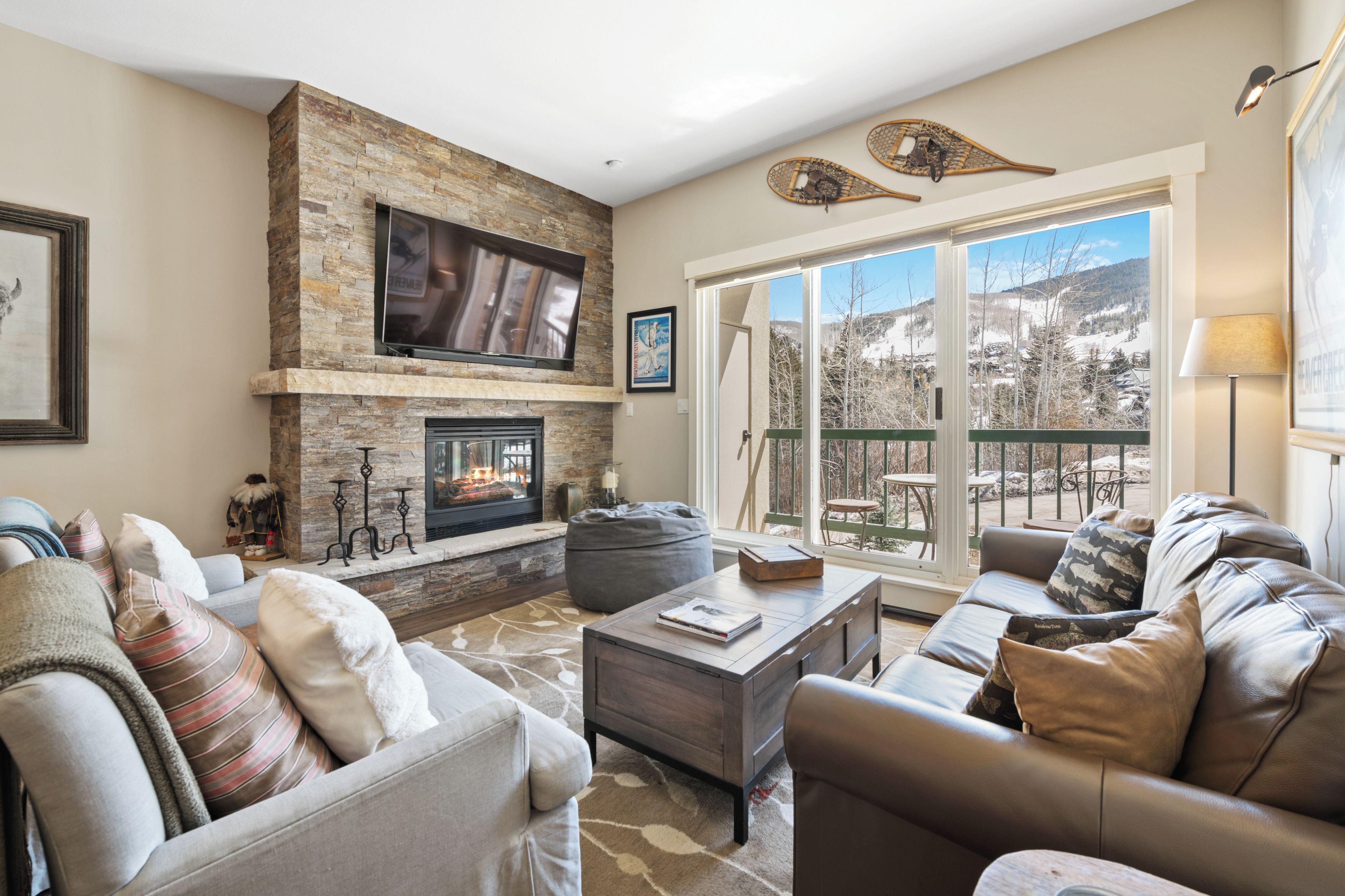 A living room with a fireplace, leather sofas, a flat-screen TV, and a large window with a view of a snowy landscape.